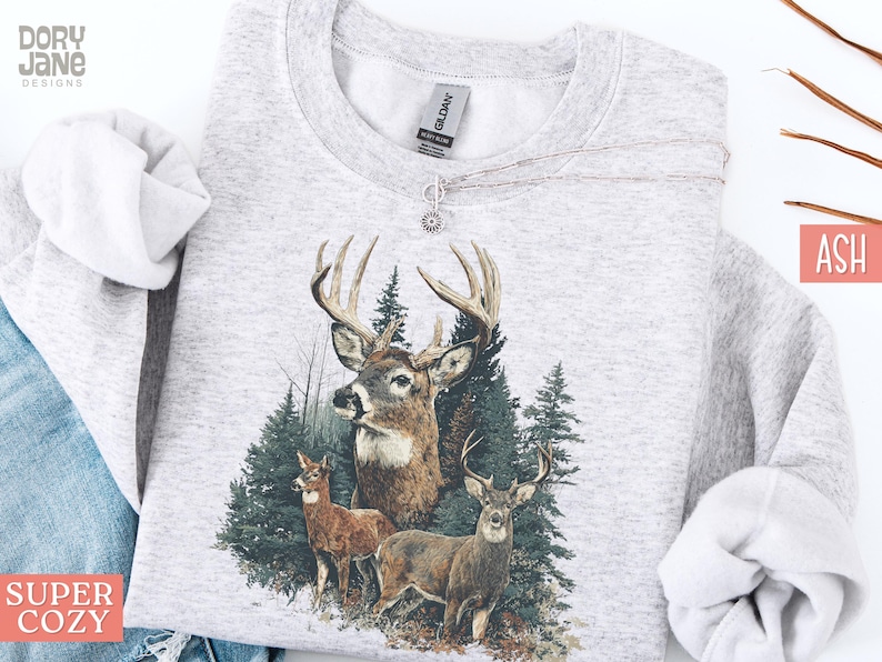 Pu&ograve; includere: Felpa color cenere con una grafica dettagliata di tre cervi in un ambiente forestale. Il design include un grande cervo con corna, due cervi pi&ugrave; piccoli e alberi sempreverdi. Le parole "SUPER COZY" sono stampate in basso a sinistra.