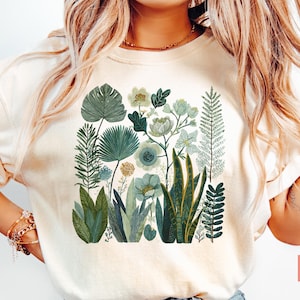 May include: Cream-coloured t-shirt with a botanical illustration. The design features various green and blue-green plants and flowers, including large leaves, ferns, and floral arrangements. The text "Dory Jane Designs" is visible at the bottom left.