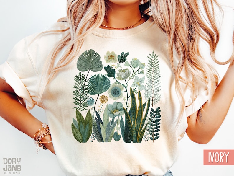 May include: Cream-coloured t-shirt with a botanical illustration. The design features various green and blue-green plants and flowers, including large leaves, ferns, and floral arrangements. The text "Dory Jane Designs" is visible at the bottom left.