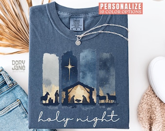Puede incluir: Una camiseta azul marino con un diseño de acuarela de una escena de la natividad y el texto "holy night". El diseño incluye una estrella, un establo y siluetas de figuras y animales. Un collar plateado con un colgante está sobre la camiseta.