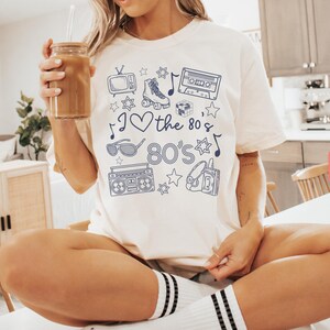 Puede incluir: Una camiseta blanca con un dise&ntilde;o gr&aacute;fico azul que presenta una cinta de casete, patines, gafas de sol, una radiograbadora y el texto "I love the 80's".