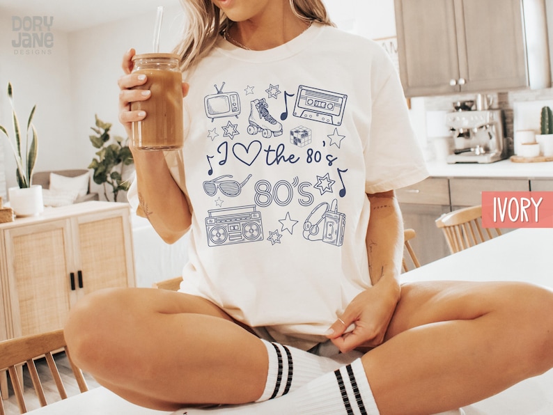 Puede incluir: Una camiseta blanca con un dise&ntilde;o gr&aacute;fico azul que presenta una cinta de casete, patines, gafas de sol, una radiograbadora y el texto "I love the 80's".