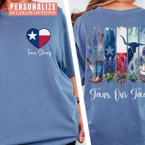 May include: Blue t-shirt with a Texas-themed design. The front features a heart with the Texas flag and the text "Texas Strong." The back displays a Texas flag design with boots, flowers, and a longhorn, with the text "Texas, Our Texas."
