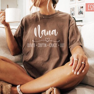 May include: A brown t-shirt with the word "Nana" and a heart design. Below the word "Nana" are the names "Oliver, Griffin, Gracie, Lily".