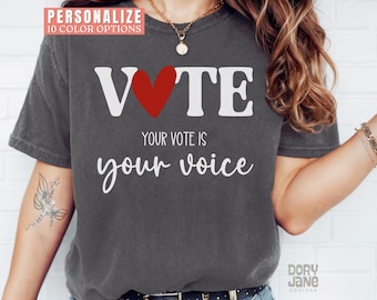 May include: A dark gray t-shirt with the word "VOTE" in white letters and a red heart in the middle. The text "Your vote is your voice" is printed below the word "VOTE".