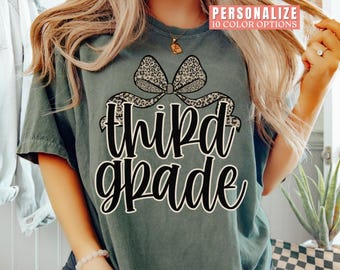 May include: A sage green t-shirt with a large bow design featuring a leopard print pattern. The words "third grade" are written in a bold, cursive font below the bow. The shirt is a comfortable, casual style.