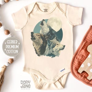 May include: Cream-colored baby onesie featuring a graphic of three wolves howling at a full moon. The design is set against a teal circle. The onesie is made of Gerber Premium Cotton. A wooden giraffe toy and a bib are next to the onesie.