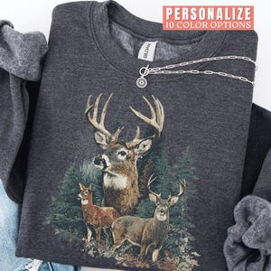 Pu&ograve; includere: Felpa grigio scuro con una grafica dettagliata di tre cervi in un ambiente forestale. Il design include un grande cervo con corna prominenti e due cervi pi&ugrave; piccoli. Il testo "PERSONALIZE 10 COLOR OPTIONS" &egrave; visibile.