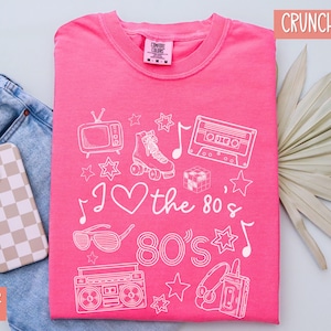 Puede incluir: Camiseta rosa con un dise&ntilde;o gr&aacute;fico blanco con iconos de los 80 como una radiograbadora, una cinta de casete, patines y gafas de sol. El texto "I love the 80's 80's" tambi&eacute;n est&aacute; incluido en el dise&ntilde;o.