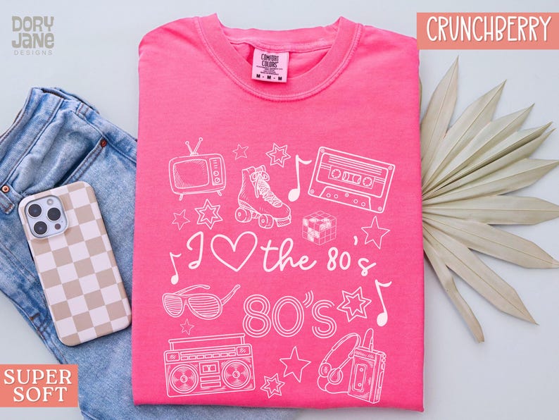 Puede incluir: Camiseta rosa con un dise&ntilde;o gr&aacute;fico blanco con iconos de los 80 como una radiograbadora, una cinta de casete, patines y gafas de sol. El texto "I love the 80's 80's" tambi&eacute;n est&aacute; incluido en el dise&ntilde;o.