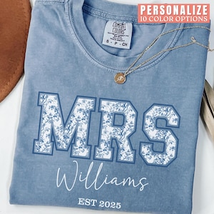 May include: A light blue t-shirt with a white floral design that says "MRS Williams EST 2025".