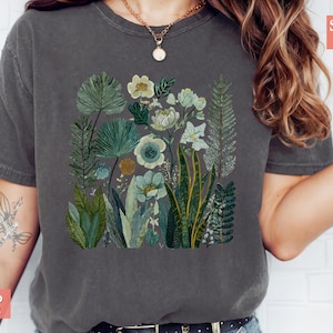 May include: Dark grey t-shirt with a botanical illustration of plants and flowers in green, white, and blue. The design features detailed leaves and floral elements, creating a natural, artistic aesthetic. The shirt is labelled "SUPER SOFT" and "PEPPER".