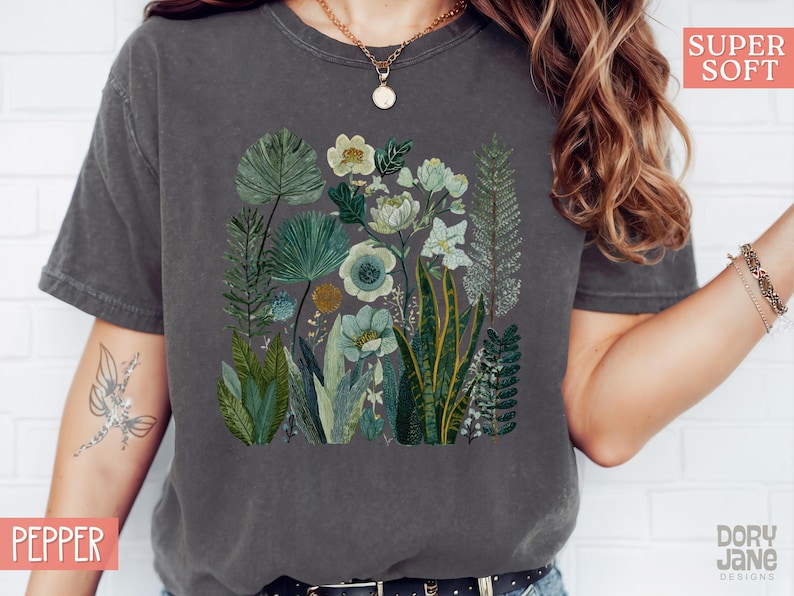 May include: Dark grey t-shirt with a botanical illustration of plants and flowers in green, white, and blue. The design features detailed leaves and floral elements, creating a natural, artistic aesthetic. The shirt is labelled "SUPER SOFT" and "PEPPER".