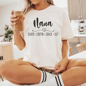 May include: White t-shirt with the text "Nana" in black script font with a small heart above the word. Below the word "Nana" is the text "Oliver - Griffin - Gracie - Lily" in black.