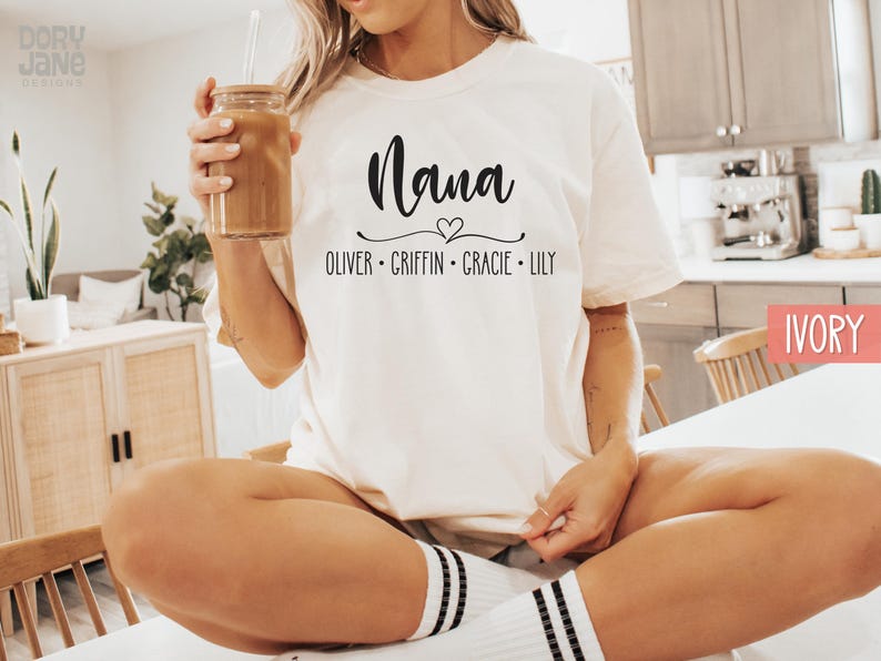 May include: White t-shirt with the text "Nana" in black script font with a small heart above the word. Below the word "Nana" is the text "Oliver - Griffin - Gracie - Lily" in black.