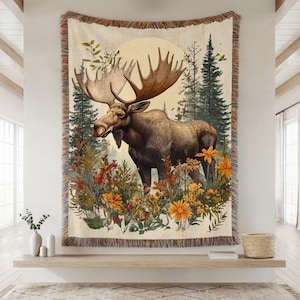 May include: A woven throw blanket featuring a large brown moose standing in a forest with tall pine trees and a pale moon in the background. The foreground is filled with colourful wildflowers in shades of orange and yellow, with fringed edges.