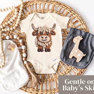Pode incluir: Um body de beb&eacute; creme com um design bordado de vaca Highland. A imagem inclui um pano de musselina cinzento, um brinquedo de girafa de madeira e uma grinalda de contas, tudo disposto numa bandeja de tecido. O texto diz "Gentle on Baby's Skin".