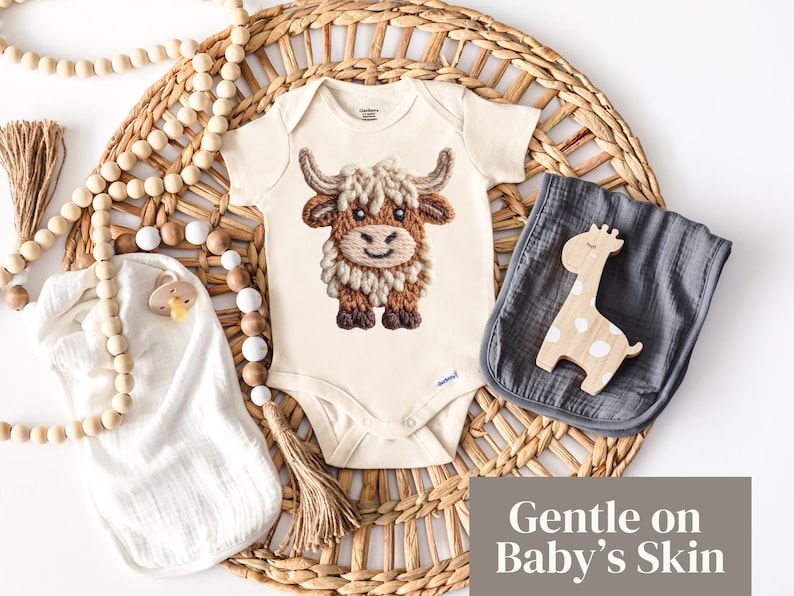 Pode incluir: Um body de beb&eacute; creme com um design bordado de vaca Highland. A imagem inclui um pano de musselina cinzento, um brinquedo de girafa de madeira e uma grinalda de contas, tudo disposto numa bandeja de tecido. O texto diz "Gentle on Baby's Skin".