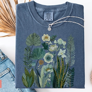 May include: A denim blue Comfort Colors t-shirt with a botanical illustration of flowers and foliage in green, white, and yellow. A silver necklace with a circular pendant rests on the shirt. The word "DENIM" is visible.