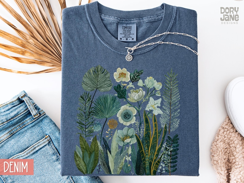 May include: A denim blue Comfort Colors t-shirt with a botanical illustration of flowers and foliage in green, white, and yellow. A silver necklace with a circular pendant rests on the shirt. The word "DENIM" is visible.