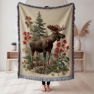 May include: Beige woven throw blanket featuring a vintage illustration of a moose with large antlers, surrounded by red flowers and a forest scene. The blanket has a fringed edge in shades of blue and beige.