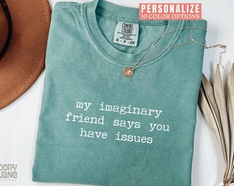 May include: A seafoam green t-shirt with the text "my imaginary friend says you have issues" in white. A gold-colored necklace with a small pendant rests on the shirt. The shirt has a Comfort Colors tag.