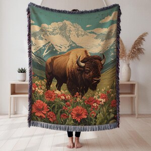 May include: Woven throw blanket featuring a bison in a field of red and white flowers, with a mountain range backdrop. The blanket has a fringed edge and a vintage-inspired design. The color palette includes shades of brown, green, and blue.