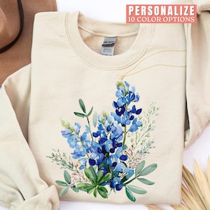 May include: Cream-colored sweatshirt featuring a watercolor design of bluebonnet flowers with green leaves and pink accents. The text "PERSONALIZE 10 COLOR OPTIONS" is at the top. A gold necklace is draped across the sweatshirt.