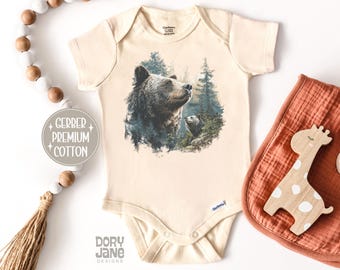 May include: Cream-colored baby onesie featuring a detailed illustration of a bear and cub in a forest setting. The onesie has short sleeves and snap closures. The text "Gerber Premium Cotton" is visible. A bib and wooden giraffe are also present.