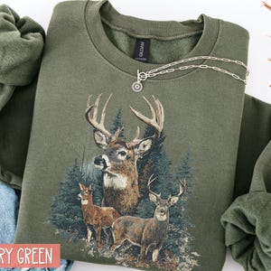 Pu&ograve; includere: Felpa verde militare con una grafica dettagliata di tre cervi in una foresta. Il design include un grande cervo con corna e due cervi pi&ugrave; piccoli. Una collana d'argento &egrave; drappeggiata sulla felpa.