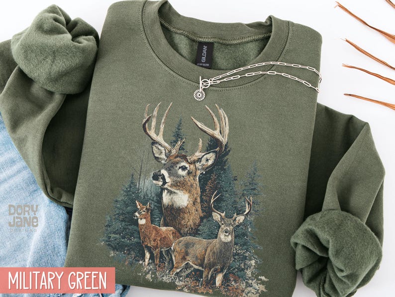 Pu&ograve; includere: Felpa verde militare con una grafica dettagliata di tre cervi in una foresta. Il design include un grande cervo con corna e due cervi pi&ugrave; piccoli. Una collana d'argento &egrave; drappeggiata sulla felpa.
