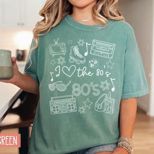 Puede incluir: Una camiseta verde claro con un dise&ntilde;o gr&aacute;fico blanco que presenta iconos de los 80 como una radiocassette, patines, una cinta de casete y gafas de sol. El texto de la camiseta dice "I &hearts; the 80's 80's".