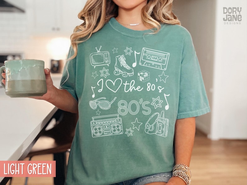 Puede incluir: Una camiseta verde claro con un dise&ntilde;o gr&aacute;fico blanco que presenta iconos de los 80 como una radiocassette, patines, una cinta de casete y gafas de sol. El texto de la camiseta dice "I &hearts; the 80's 80's".