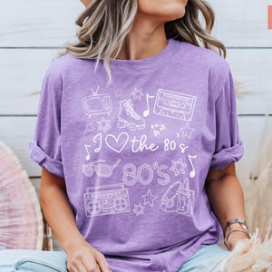 Puede incluir: Una camiseta morada con un dise&ntilde;o gr&aacute;fico blanco que presenta el texto "I love the 80's 80's" y varios iconos de los 80, incluyendo patines, una cinta de casete, una radiograbadora y gafas de sol.