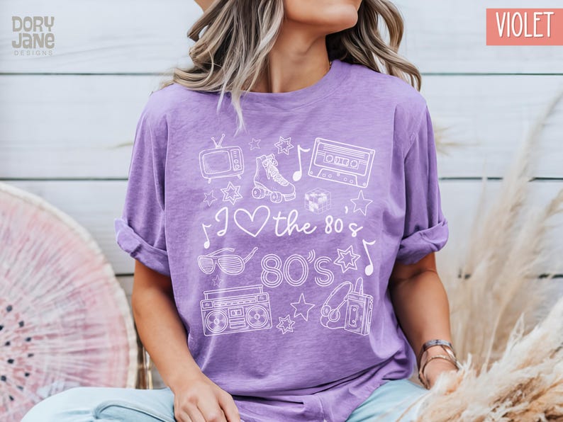 Puede incluir: Una camiseta morada con un dise&ntilde;o gr&aacute;fico blanco que presenta el texto "I love the 80's 80's" y varios iconos de los 80, incluyendo patines, una cinta de casete, una radiograbadora y gafas de sol.