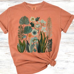 May include: Coral-coloured t-shirt with a botanical illustration of diverse plants and flowers in green, blue, and white. The design features detailed leaves and floral elements, creating a natural theme. The shirt has short sleeves and a crew neck.