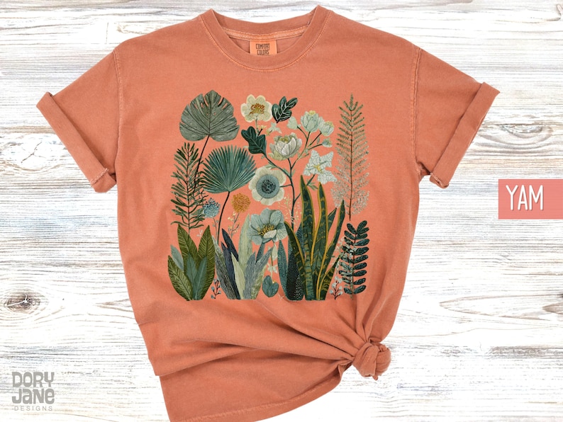 May include: Coral-coloured t-shirt with a botanical illustration of diverse plants and flowers in green, blue, and white. The design features detailed leaves and floral elements, creating a natural theme. The shirt has short sleeves and a crew neck.