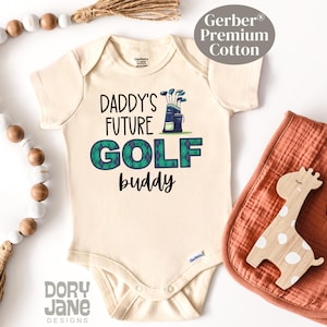May include: A cream-colored baby onesie with short sleeves and the text "Daddy's Future Golf Buddy" in black. A green and blue plaid golf bag with clubs is pictured above the text.