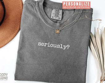 May include: A gray t-shirt with the word "seriously?" printed in white. A gold necklace with a pendant rests on the shirt. The shirt has a Comfort Colors tag. A brown hat and dried leaves are in the background.
