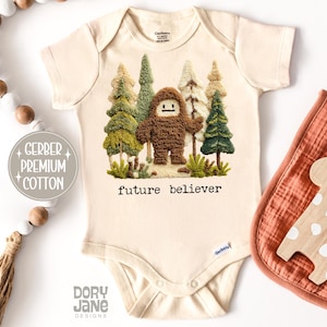 May include: Cream-colored baby onesie with a Bigfoot design. The graphic features a brown Bigfoot figure standing in front of green and tan trees. The text "future believer" is printed below the image. The onesie is made of Gerber premium cotton.