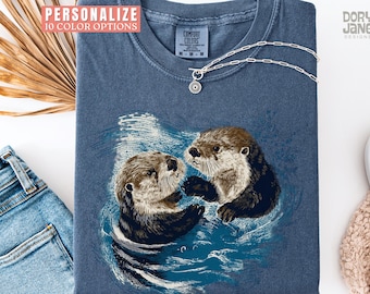 May include: Blue Comfort Colors t-shirt featuring an illustration of two otters in water. The shirt has the text "PERSONALIZE 10 COLOR OPTIONS". A silver necklace with a sun pendant is on the shirt.