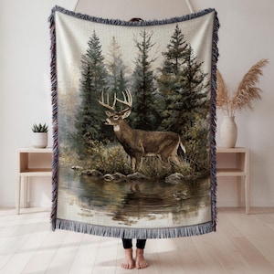 May include: Woven throw blanket featuring a detailed illustration of a deer standing near a body of water in a forest. The blanket has a fringed edge with shades of blue, brown, and green. The deer has large antlers and is brown with white accents.