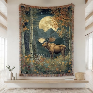 May include: A woven tapestry blanket featuring a moose standing in a meadow of colourful wildflowers. The scene includes tall birch trees, a dark forest, and a large full moon set against a starry night sky. The edges have a multicoloured fringe.