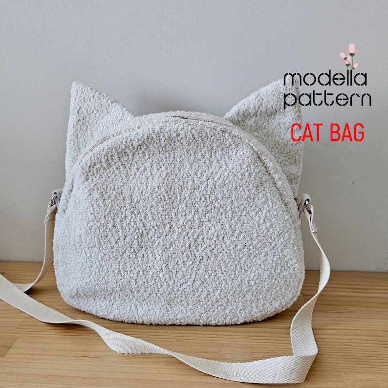 Op de afbeelding: Een witte, pluche, kattenvormige schoudertas met een witte band. De tas heeft een ritssluiting en de tekst "modella pattern CAT BAG" is op de tas gedrukt.