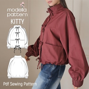 Può includere: Una giacca rosso scuro con colletto alto, maniche lunghe con polsini arricciati e chiusura frontale con molteplici laccetti da annodare. L'immagine include il testo modella pattern KITTY e Pdf Sewing Pattern con illustrazioni a linee.