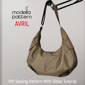 May include: A brown, fabric handbag with a long strap hanging from a black metal arm. The bag has a zipper closure and a small pocket on the side. The text "modela pattern AVRIL" is printed on a white background above the bag.