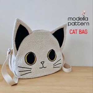 Op de afbeelding: Een witte pluche crossbody tas in de vorm van een kat met zwarte oren, gele ogen en een zwarte geborduurde neus en snorharen. De tas heeft een witte band en de tekst "modella pattern CAT BAG" is gedrukt op een witte achtergrond.