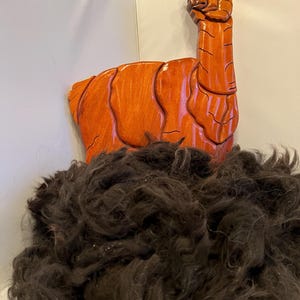 May include: A carved wooden llama sculpture in shades of orange, with detailed features. Below the sculpture is a pile of dark brown alpaca fiber, ready for crafting. The llama is mounted on a white wall.
