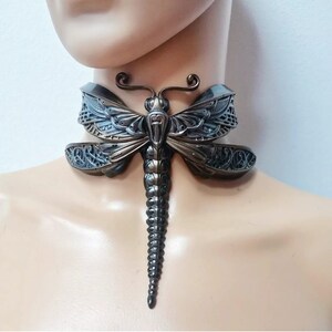 Può includere: Una collana girocollo a forma di libellula color bronzo. La collana presenta un design dettagliato di libellula con intricati motivi alari e un corpo segmentato. Il girocollo è indossato intorno al collo.