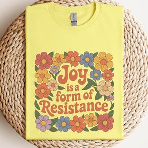 May include: A yellow t-shirt with the text "Joy is a form of Resistance" in orange lettering, surrounded by a floral design in shades of orange, yellow, blue, and purple. The shirt is displayed on a woven, circular surface.
